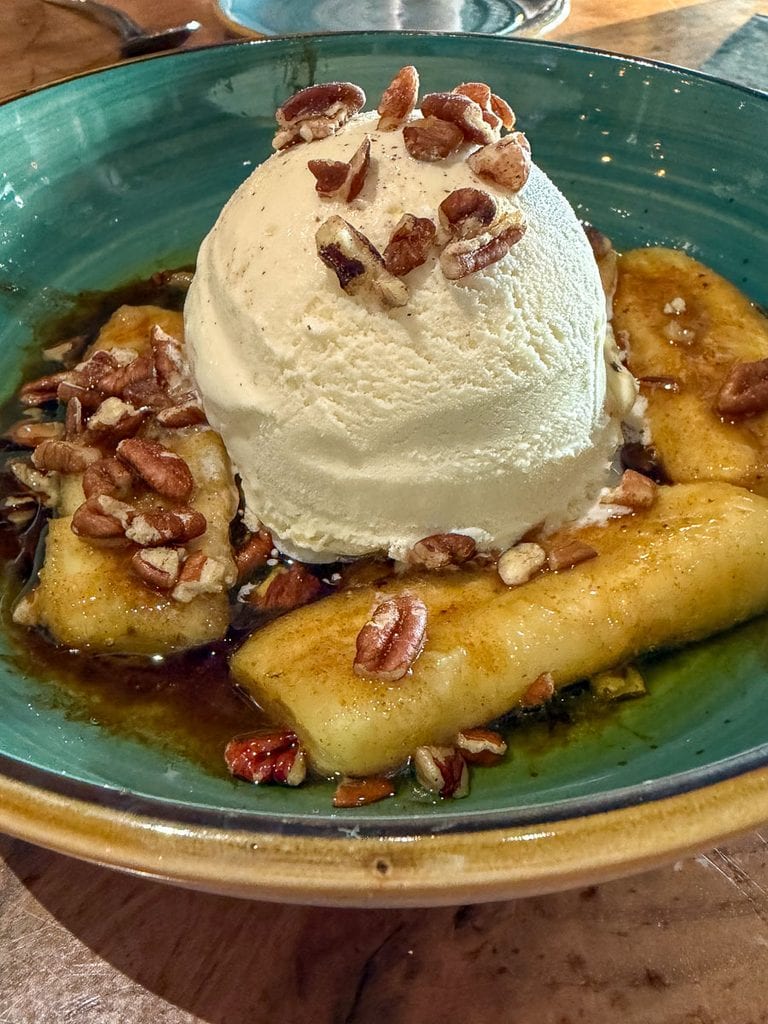 Vanilla ice cream topped with pecans, served with caramelized banana slices in a green bowl on a wooden table—a sweet treat reminiscent of dessert after a multigenerational trip to Ann Arbor.