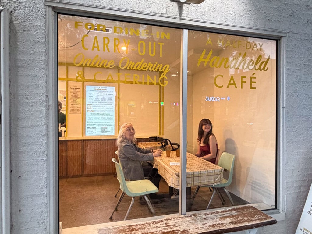 Two people sit at a table with a checkered tablecloth inside a café, seen through a window with yellow lettering advertising dine-in, carry-out, and catering. The café is warmly lit, menus are visible on the wall, and the cozy atmosphere makes it an inviting stop during a multigenerational trip to Ann Arbor.