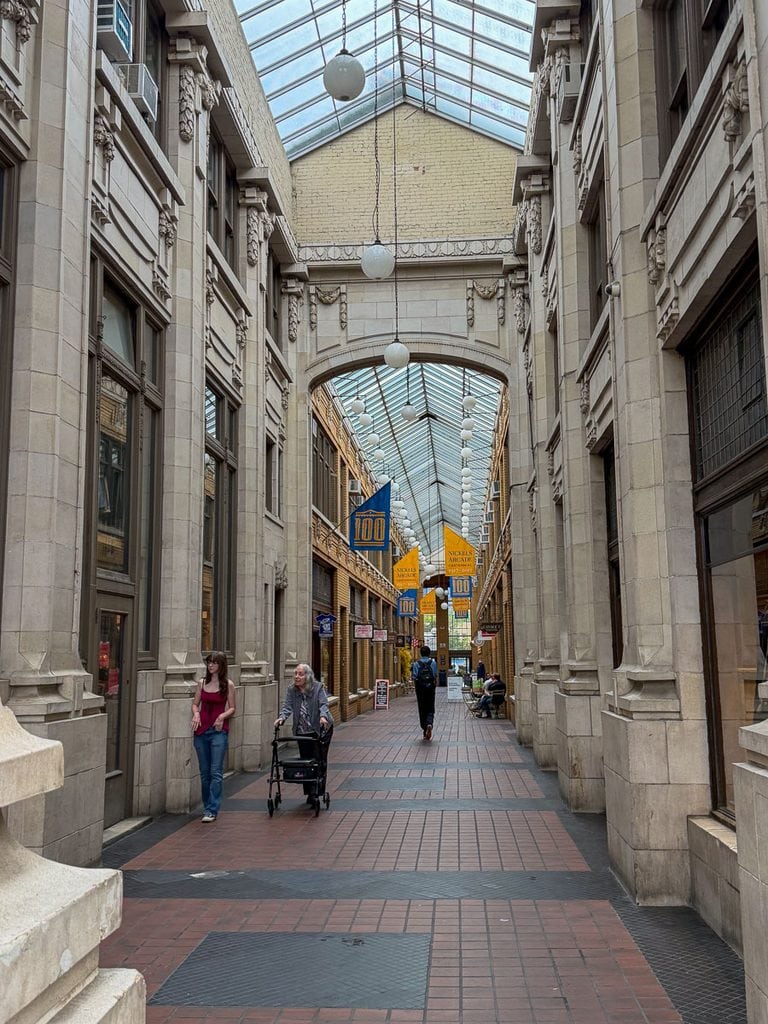 A covered shopping arcade with tall arched windows, ornate stone architecture, hanging lights, and people walking or sitting makes a charming stop during a multigenerational trip to Ann Arbor. Blue and yellow banners hang from the ceiling, and the floor is tiled in red and brown.